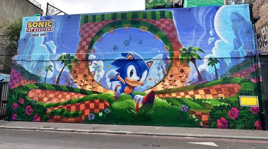 The image depicts a vibrant mural painted on a brick wall, featuring Sonic the Hedgehog in a dynamic pose. Sonic is depicted in a blue and white color scheme, with his arms raised, as if in motion. The mural is set against a backdrop of a green landscape with palm trees, a checkered pattern, and a blue sky with clouds.