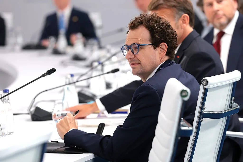 The image shows a man seated at a table in a conference room, wearing a dark blue suit and glasses. He is holding a pen and appears to be engaged in a discussion or presentation. The table is covered with a white tablecloth and features microphones and water bottles. In the background, other individuals are seated at t