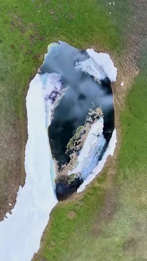 The image presents an aerial view of a small, frozen lake nestled in a grassy area. The lake's surface is partially covered in ice, with some areas showing a mix of white and blue hues. The surrounding grassy area is a vibrant green, contrasting with the white and blue ice. The frozen lake is surrounded by a mix of bro