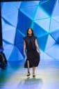 The image shows a person with a prosthetic leg walking on a runway. The individual is wearing a dark blue dress and a dark blue vest over a black top. The prosthetic leg is visible and extends from the person's right foot to their left foot. The runway is set against a backdrop of a blue geometric pattern. The person's