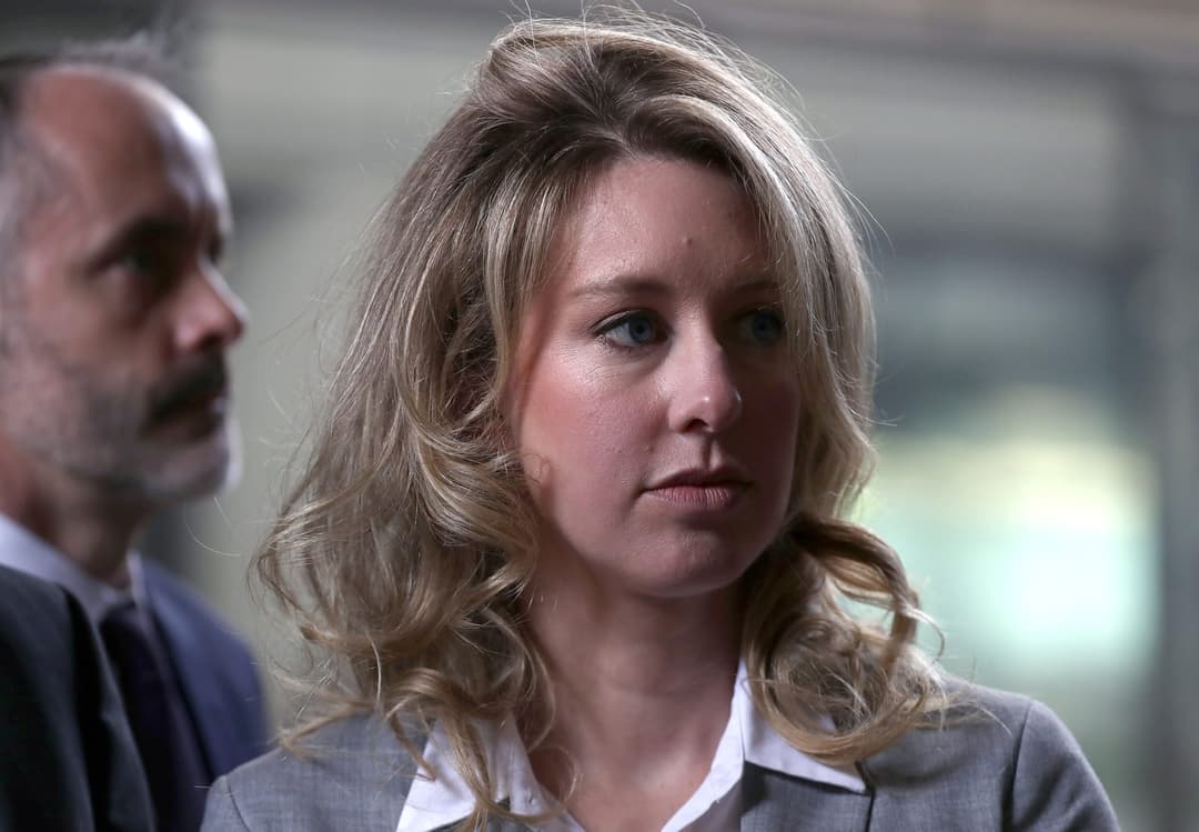 The image shows a woman with blonde hair, styled in loose curls, wearing a gray blazer over a white collared shirt. She is looking off to the side with a serious expression. Behind her, a man with a beard and mustache is partially visible, wearing a dark suit and tie. The background is blurred, suggesting an indoor set