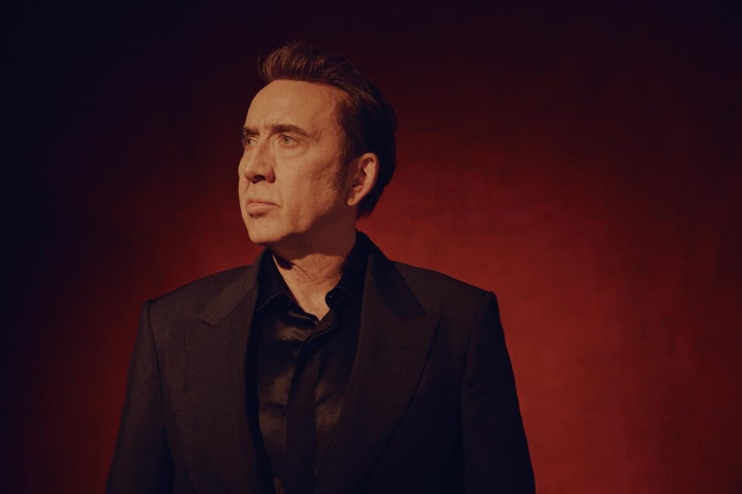 The image shows a man standing against a dark red background. He is dressed in a black suit and a black shirt, and his gaze is directed off to the side. His hair is styled in a neat, combed-back manner. The image is captured from a slightly low angle, emphasizing the man's stature. The man's expression is neutral, and