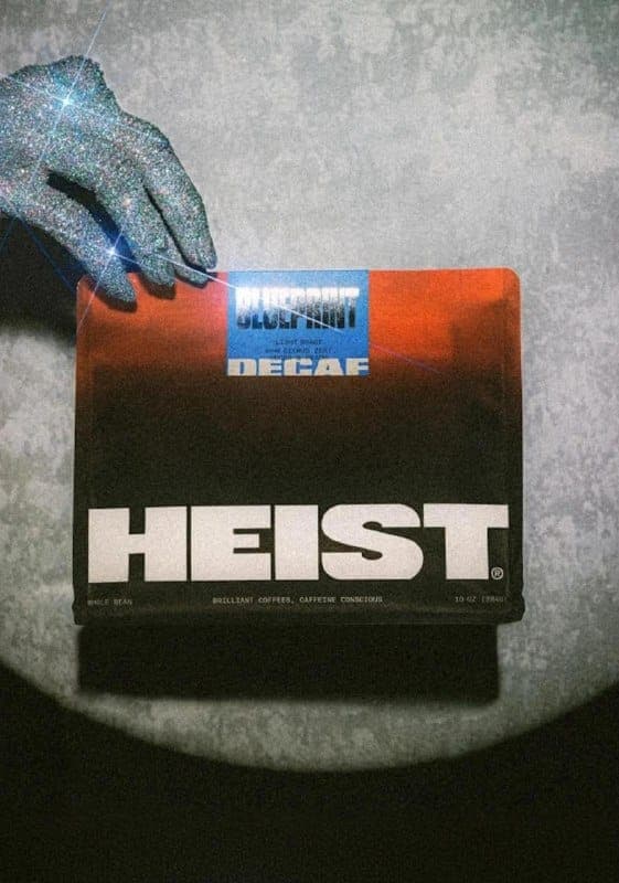 The image shows a hand adorned with a glittery, silver-colored glove resting on a black surface. The hand is positioned over a rectangular box of "Heist" coffee, which is the central focus of the image. The box is primarily black with a gradient of orange and red colors, and features a blue label with the words "Bluepr