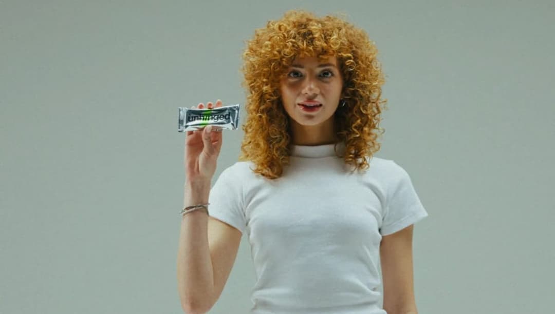 The image shows a young woman with curly blonde hair standing against a light gray background. She is wearing a white t-shirt and has a surprised or shocked expression on her face. In her right hand, she holds a bar of soap with a black wrapper and a green label that reads "Uniqed".