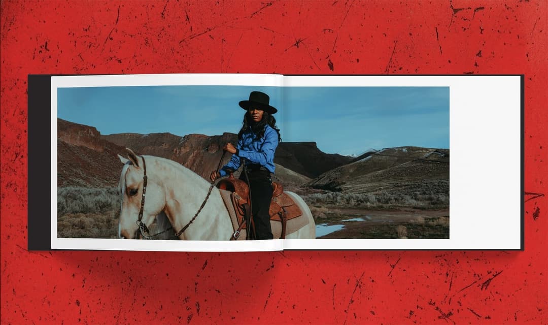 The image shows a woman dressed in a blue shirt and black hat, riding a white horse in a desert landscape. The horse is adorned with a brown saddle and bridle. The woman is holding a riding crop in her left hand. The landscape features a mix of brown and green vegetation, with a clear blue sky overhead. The scene is ca