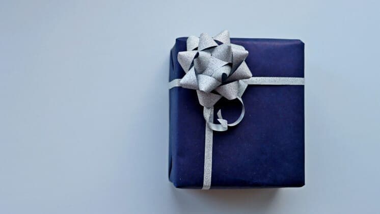 The image shows a single, square gift box wrapped in dark blue paper. The box is adorned with a silver bow on top, which is tied in a bow with a silver ribbon. The gift box is placed on a light blue background, which provides a neutral backdrop that allows the box and its decorations to stand out.