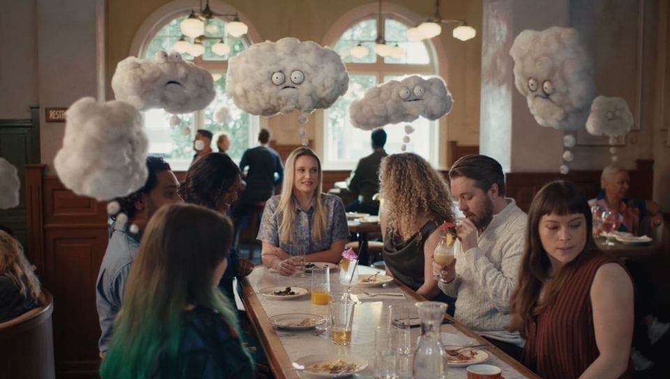 The image shows a group of people gathered around a long, white table in a restaurant. The table is set with plates, glasses, and cutlery, and some people are holding drinks. The restaurant has a classic, possibly old-fashioned, aesthetic with wooden paneling on the walls and arched windows. The ceiling features a deco
