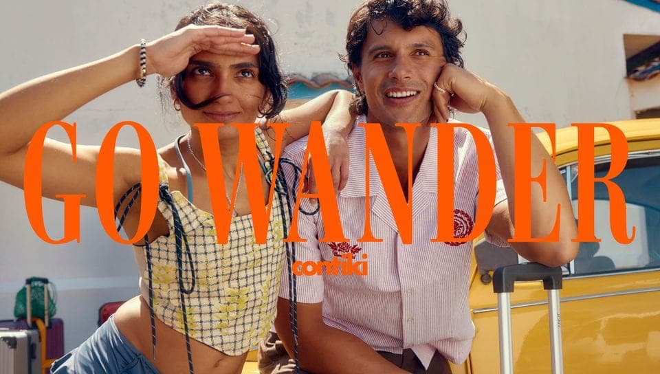 A young man and woman stand in front of a white wall and yellow vehicle, the woman shading her eyes and the man smiling with his hand on his cheek, with large orange serif text reading "GO WANDER" and the Contiki logo across the image.