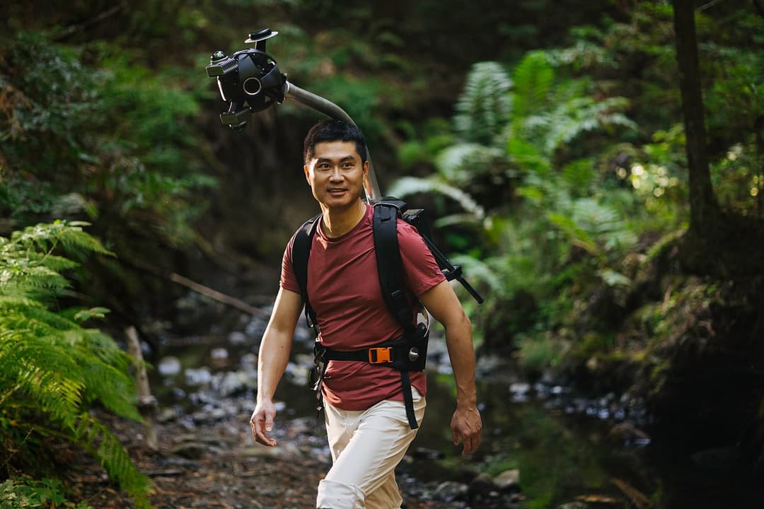 A man is walking along a narrow stream in a lush forest. He is wearing a vibrant red t-shirt and white pants, and is carrying a black backpack with a camera attached to the top. The camera is equipped with a large lens and a sturdy black tripod. The stream is surrounded by a variety of green ferns and trees, creating a