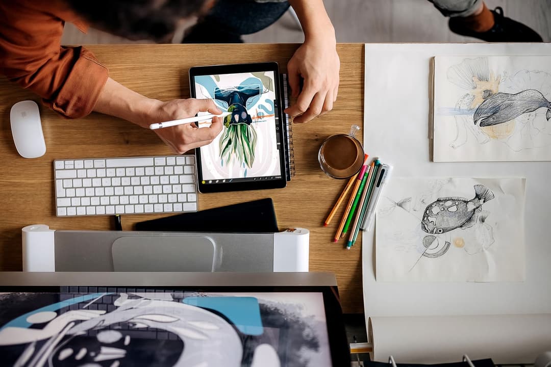 Overhead view of a wooden desk where a person draws a colorful illustration on a tablet with a stylus, next to a keyboard, mouse, coffee cup, colored pencils, and pencil sketches of fish and whales on paper.