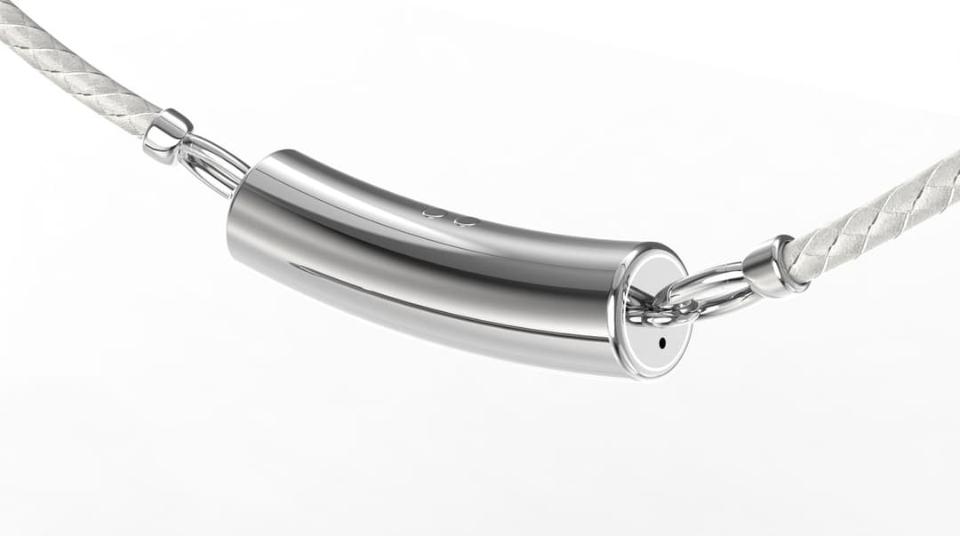 The image shows a silver-colored necklace with a unique cylindrical pendant. The pendant is attached to a silver-colored metal loop, which is connected to a silver-colored metal chain. The chain is made of a white, braided or twisted material. The pendant is positioned slightly to the left of the center of the image. T