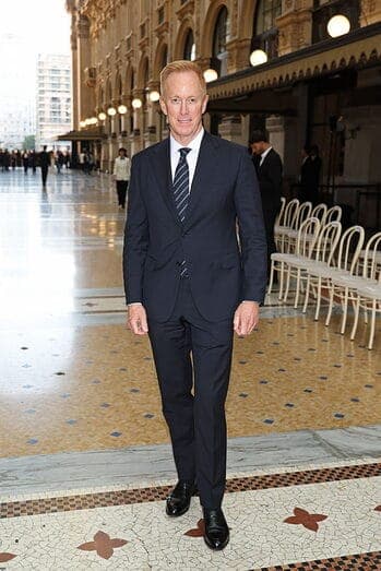 The image shows a man in a dark blue suit and tie standing in the center of a grand, ornate interior space. The man is wearing black dress shoes and has light brown or blond hair. The interior features a large, ornate building with arched windows and a decorative awning, and a tiled floor with a mosaic pattern of brown