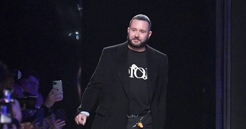 The image shows a man walking down a dark runway, likely during a fashion show. He is wearing a black blazer over a black t-shirt with a white logo and black pants. He has short, dark hair and a beard. The runway is dark, and the background is mostly dark, with a few indistinct lights. In the foreground, a group of peo