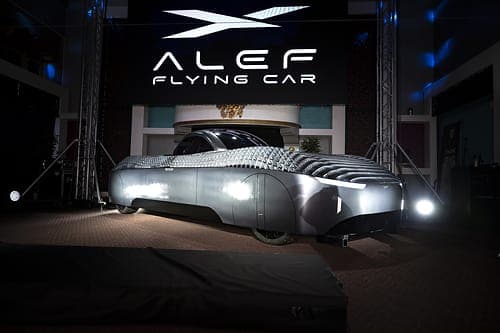 A silver, textured flying-car prototype stands on a dark stage in front of a backdrop with the logo "ALEF FLYING CAR" and an angled wing-like emblem, lit by spotlights. The vehicle has a sleek, panelled body with visible wheels.