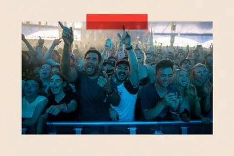 The image captures a lively concert scene with a large crowd of people enthusiastically cheering and raising their hands. The crowd is densely packed, with individuals of various ages and genders visible. The stage is brightly lit, creating a vibrant atmosphere. The perspective of the image is from behind a barrier, pr