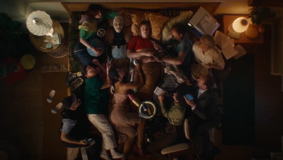 The image shows a group of people lying on a bed, with some of them appearing to be in a state of sleep or unconsciousness. The bed is covered with a variety of items, including a lamp, a globe, a book, and a small, round object that could be a toy or a decorative item. The room is dimly lit, with a lamp on a side tabl