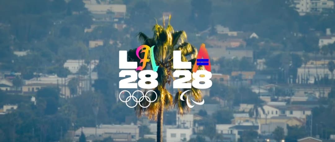 A palm tree stands in front of a hazy cityscape of low buildings and trees, with the LA28 logo overlaid in the center: "LA28" lettering with colorful script and a stylized sailboat, plus the Olympic rings and Paralympic agitos symbol.