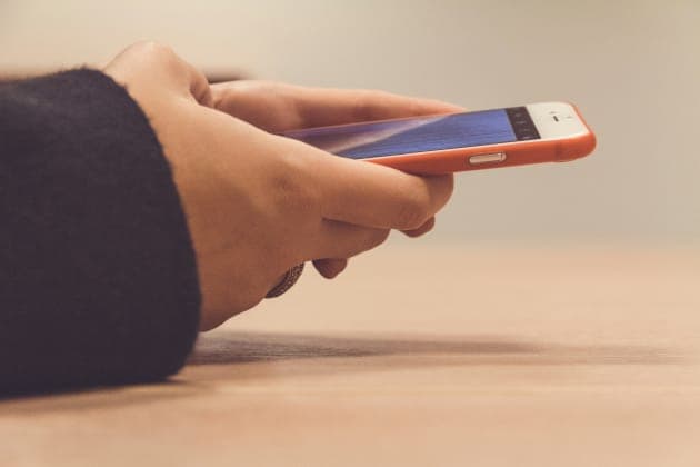 A close-up side view of a person's hands holding an orange-cased smartphone with a blue screen, with a dark sleeve on the left and a wooden surface below. No brand or readable text is visible.