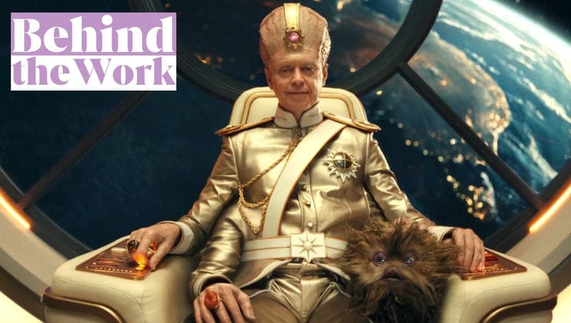 The image shows a man seated in a white, possibly leather, chair with gold accents, in a space setting. He is wearing a gold military-style uniform with a white sash and a gold medal on his chest. He is also wearing a gold, pointed-back cap with a pink gem or jewel on top. The man has a beard and is wearing a ring on h