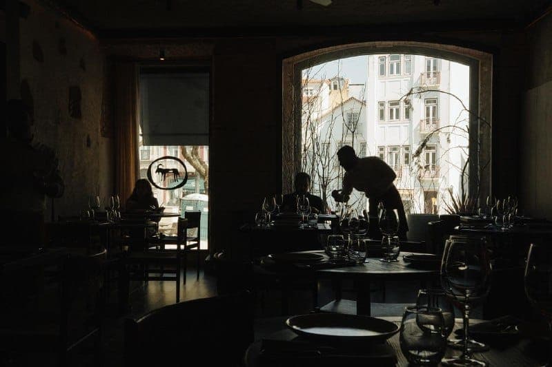 The image depicts a dimly lit restaurant with a large arched window that frames a view of a building with a white facade and a balcony. The window is covered by a white blind. The restaurant has dark wooden tables and chairs, and the tables are set with plates, glasses, and cutlery. A person is seen serving food to a c