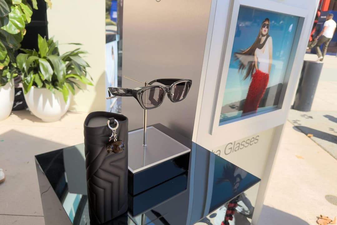 The image shows a display of sunglasses and a handbag at a store. The display is set up on a black stand with a reflective surface, positioned on a black table. The sunglasses are displayed on a stand, with a pair of black-framed sunglasses hanging from a metal rod. A black handbag with a unique geometric pattern is pl