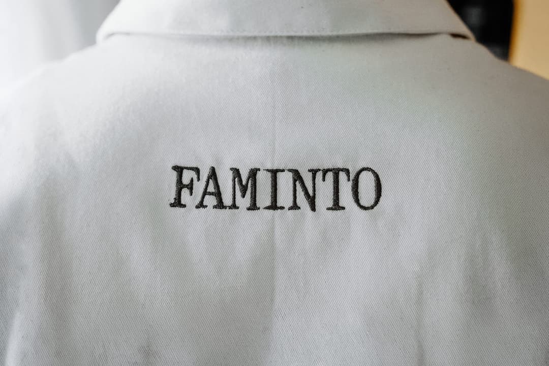 The image shows the back of a white, collared shirt. The shirt has a subtle texture, possibly a linen or cotton blend. The most prominent feature is the black embroidered text "FAMINTO" in a serif font. The embroidery is centered on the back of the shirt.
