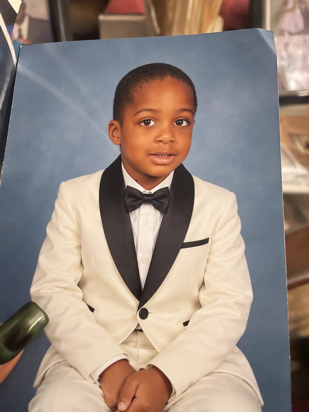 The image shows a young boy seated on a chair, wearing a cream or off-white tuxedo with a black bow tie and a black pocket square. He is looking directly at the camera with a neutral expression. The background is a solid blue color, and a portion of a dark green object is visible on the left side of the image. The boy'