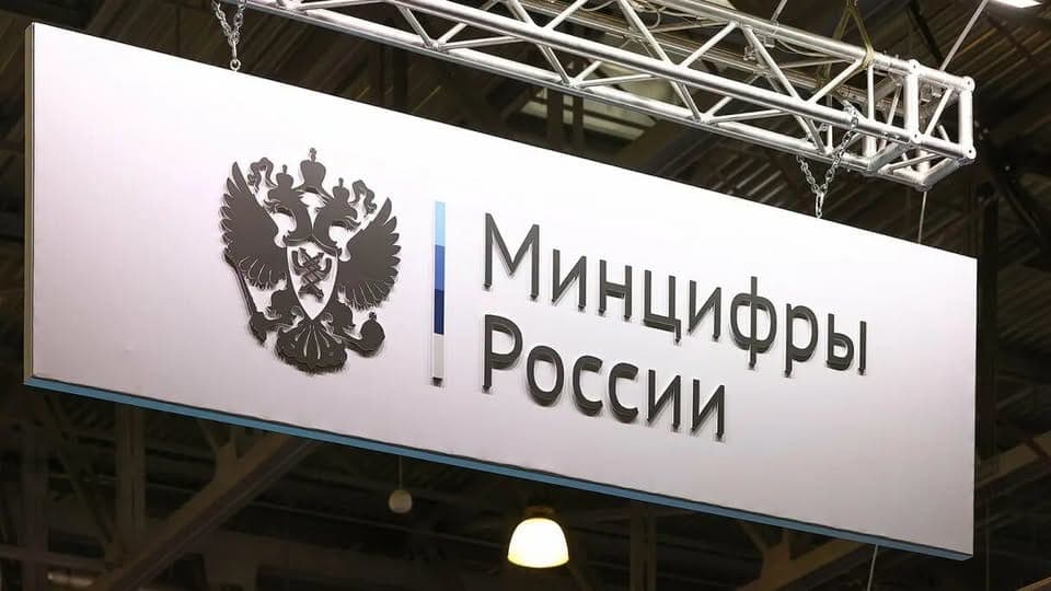 A white rectangular sign hangs from a metal truss in an industrial hall, showing a dark Russian coat of arms (double-headed eagle) and the text "Минцифры России" (Ministry of Digital Development of Russia), with a blue-and-white stripe beside the text.