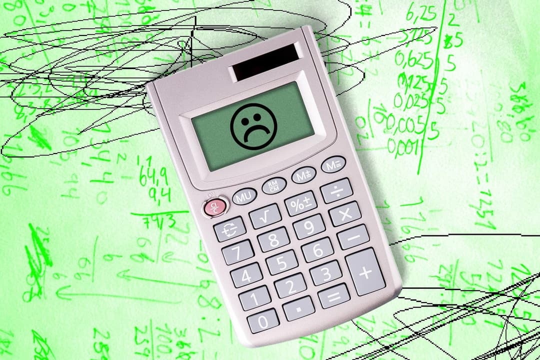 A silver pocket calculator with a green display showing a frowning face, lying on a green background covered in handwritten numbers and black scribbled loops.