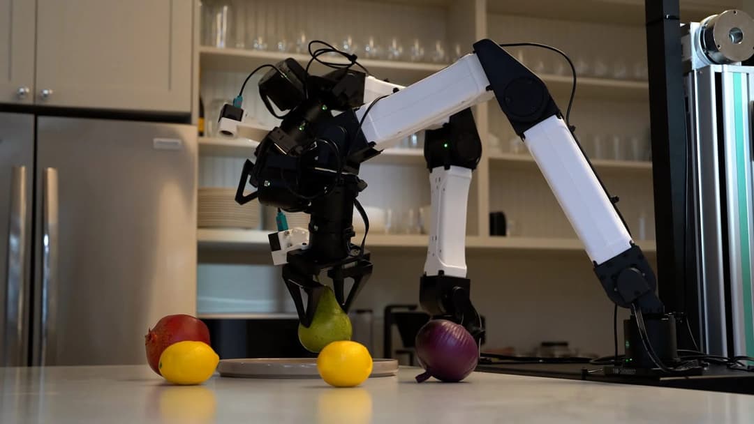 The image shows a black and white robotic arm reaching out to pick up a green pear from a white plate on a kitchen counter. The arm is positioned in the center of the image, with the pear and the plate in the foreground. The background features a kitchen with white cabinets and a stainless steel refrigerator. A shelf w