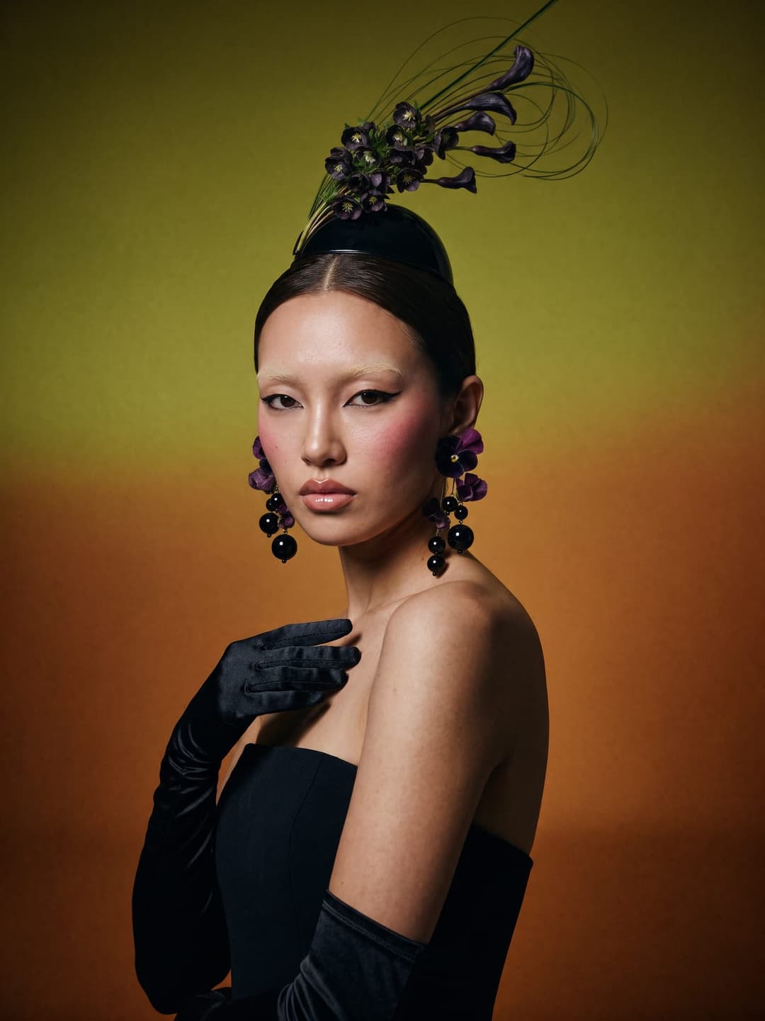 The image shows a woman as the central figure, standing against a gradient background that transitions from a dark green at the top to a warm orange at the bottom. She is dressed in a black strapless dress and black gloves, and her hair is styled in a high bun adorned with a black feather boa. She wears large, dark pur
