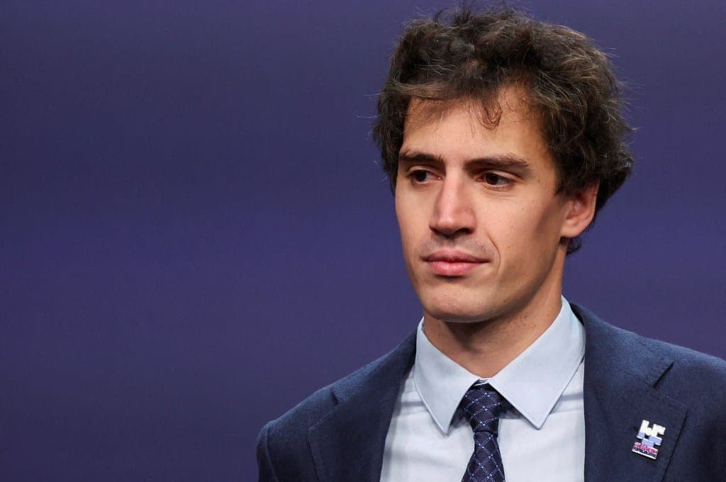 A man with curly dark hair in a navy suit, light blue collared shirt, and dark patterned tie stands against a purple backdrop, looking off to the left. A small enamel pin with blue and pink tones is on his lapel, and no visible text is legible.