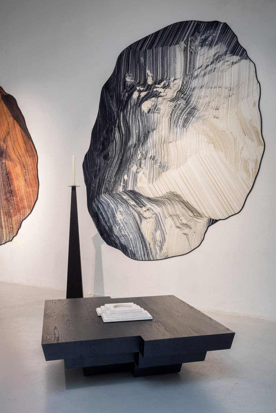 The image depicts a modern art gallery with a minimalist aesthetic. A large, abstract piece of art, primarily black and white with hints of gray and beige, dominates the space, hanging on a white wall. To the left, a tall, slender, black, triangular sculpture stands. In the center, a black, square coffee table with a w