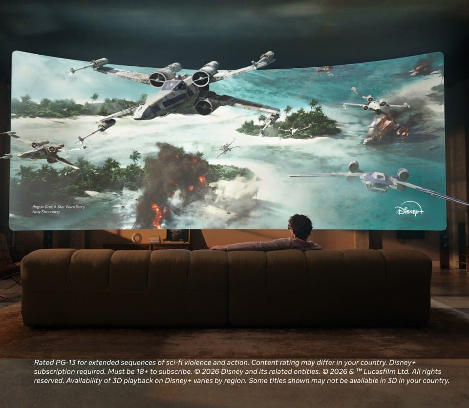 The image shows a man seated on a brown couch in a living room, watching a large curved screen displaying a scene from the Star Wars universe. The scene features a large, white X-wing fighter aircraft in flight over a tropical island with a large explosion, and other aircraft in the background. The man is wearing a lig