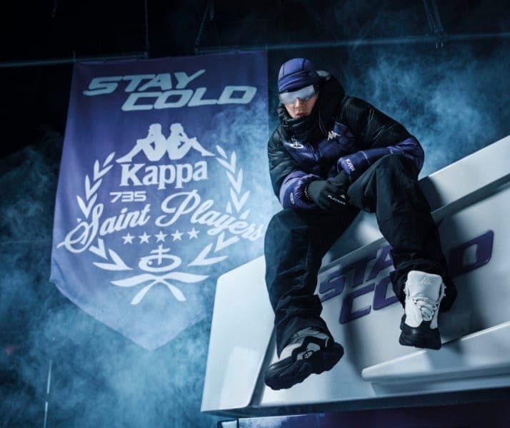 The image shows a person wearing a purple and black jacket, black pants, and a purple beanie, seated on a white bench with purple and black accents. The person is wearing white and black ski boots and is holding a pair of gloves in their lap. Behind the person, a blue banner with white text reads "STAY COLD" and featur