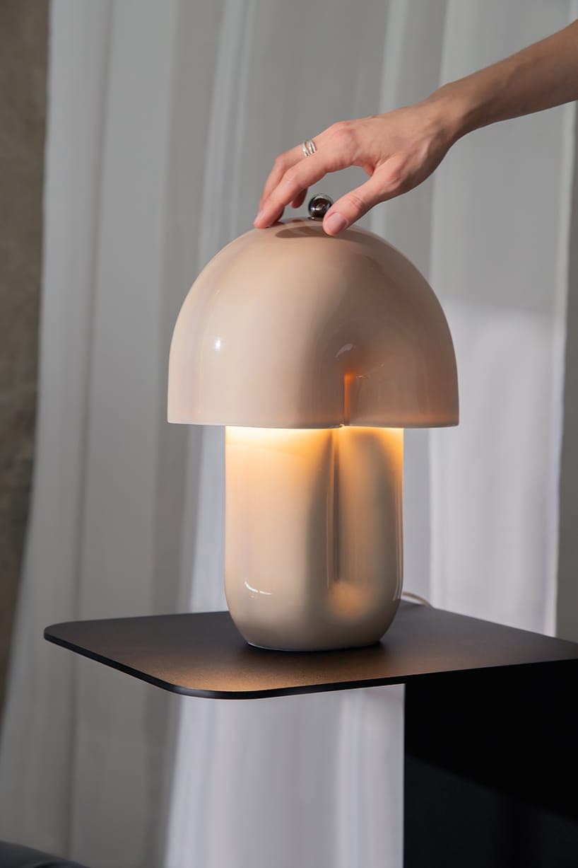 The image shows a hand gently resting on a cream-colored lamp with a unique, mushroom-like shape. The lamp has a dome-like top and a cylindrical base, and is illuminated from within, casting a warm glow. The lamp is placed on a black, square-shaped shelf, which is situated against a backdrop of white curtains. The hand