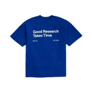 The image shows a blue t-shirt with short sleeves, laid flat against a solid green background. The t-shirt features white text that reads "Good Research Takes Time" and "Est 2016". The text is centered on the shirt.