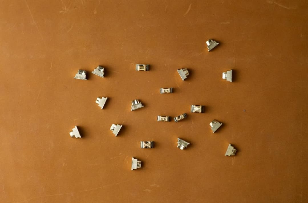 The image shows a collection of small, metallic objects scattered across a brown surface. The objects are primarily silver or light gray in color and appear to be small, possibly metallic, pieces. They are not arranged in any particular pattern, giving the image a sense of randomness. The brown surface has a slightly t