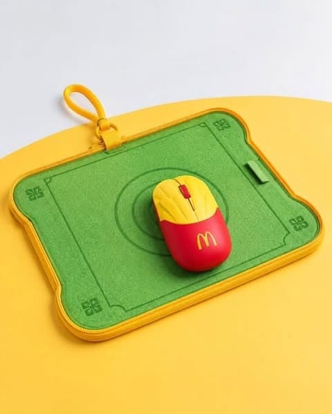 The image shows a vibrant yellow mousepad with a green border and a yellow border, featuring a McDonald's logo in red and yellow. The mousepad is resting on a yellow surface, and a red and yellow mouse with a black scroll wheel is positioned on top of it. The mousepad also has a small, yellow, looped handle attached to