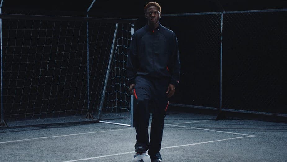 The image shows a young man standing on a dark gray or black asphalt court, possibly a soccer or basketball court. He is wearing a black tracksuit with red accents on the sleeves and pants, and white sneakers. The man is positioned in the center of the image, facing the camera with a neutral expression. The court is en