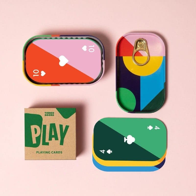 The image presents a collection of playing cards and a tin can, all set against a pink background. The playing cards are neatly arranged in a square formation, with three cards in the top row and three in the bottom row. The top row features a pink card with a red heart and the number 10, a green card with a white hear