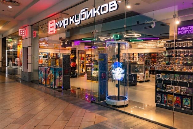 The image shows the storefront of a toy store named "��иру��ко��" (Myrkoykob) located in a mall. The store's name is prominently displayed in large, white letters above the entrance. The storefront is primarily glass, with a dark gray or black frame, and features a large, illuminated display of toys and games. The inte