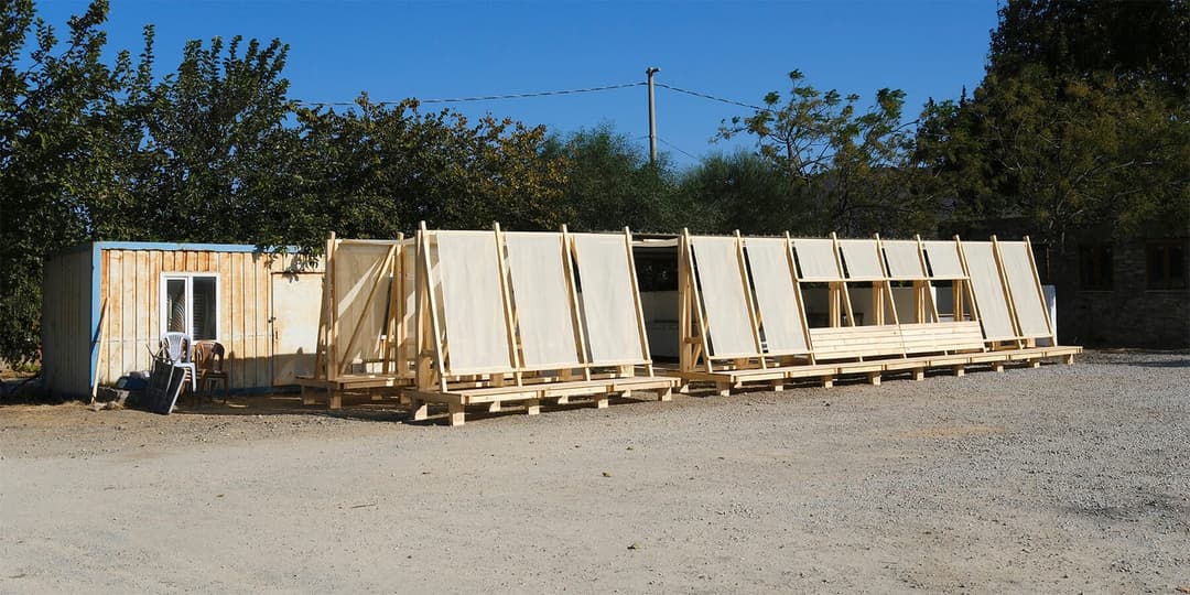 The image shows a construction site with a partially completed wooden structure. The structure is composed of several wooden planks and panels, with some sections appearing unfinished or in a state of disassembly. It is situated on a gravel or dirt surface, with a small white building with a blue roof to the left. A fe