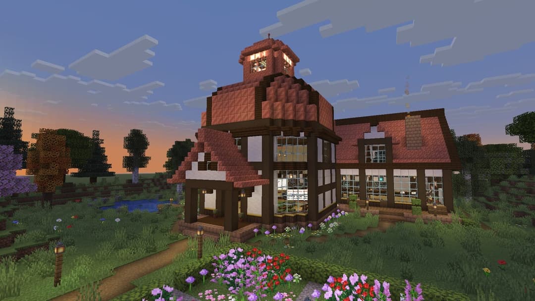 The image depicts a Minecraft house with a red tiled roof and a tower with a clock face. The house is surrounded by a lush garden filled with colorful flowers and a small pond. The house has a dark brown wooden exterior with white trim around the windows and a porch with a small railing. The sky is a gradient of blue a