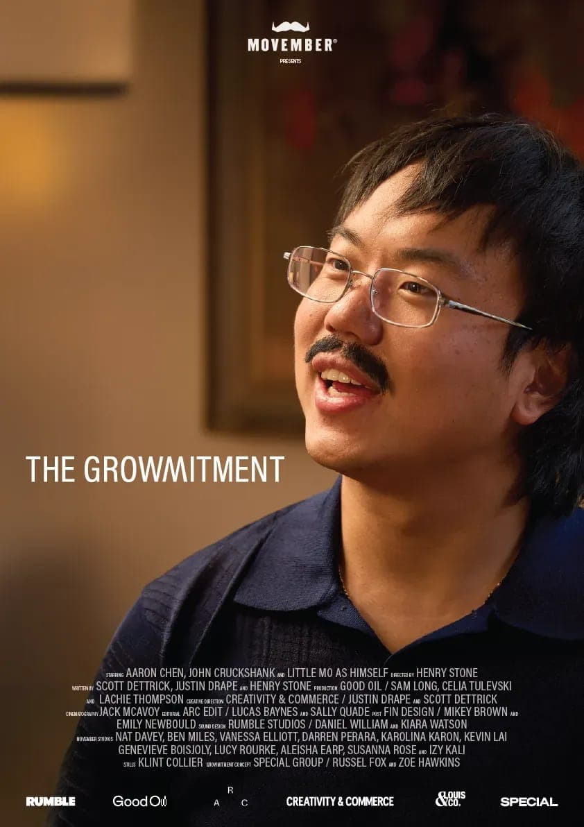 The image is a movie poster for the film "The Growthment". The poster features a man with dark hair and glasses, wearing a dark blue collared shirt, looking off to the side with a slight smile. The background is blurred, drawing focus to the man. The title of the film, "The Growthment", is prominently displayed in whit
