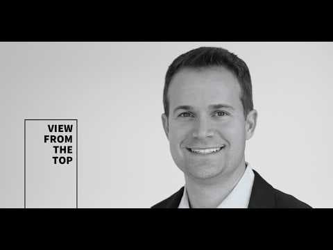 The image shows a man in a black suit and white shirt, smiling warmly at the camera. He has short, dark hair and is positioned centrally in the frame. The background is a plain white, providing a stark contrast to the man's dark suit. On the left side of the image, there is a black text box with white text that reads "