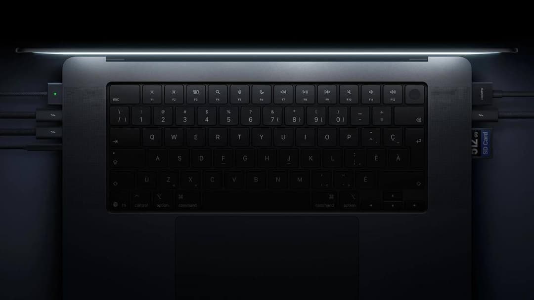 A top-down view of a dark silver laptop keyboard shows a glowing lit lid edge, a row of function keys, and a trackpad below. Cables and ports are visible on the sides, including HDMI, USB-C, and an SD card labeled "SD Card".