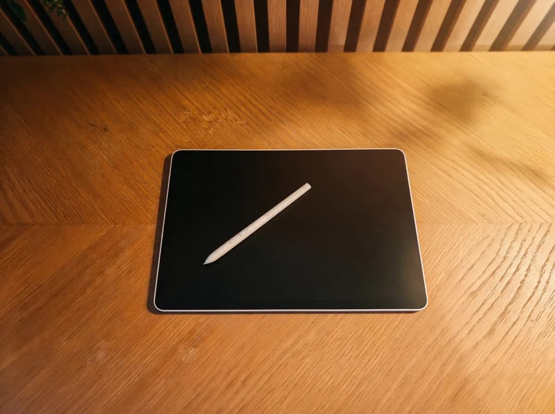 The image shows a black tablet with a white stylus resting on a wooden table. The tablet is positioned in the center of the frame, with the stylus lying diagonally across it. The wooden table has a light brown hue and a visible wood grain pattern. In the background, a wooden wall with vertical slats is visible.