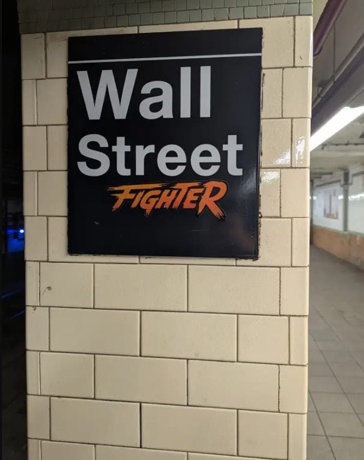 The image shows a black sign with white text that reads "Wall Street Fighter" in a bold, stylized font. The sign is affixed to a beige tiled wall, which is part of a subway station platform. The sign is slightly tilted to the left. The background of the image reveals a subway station interior, with a portion of a subwa