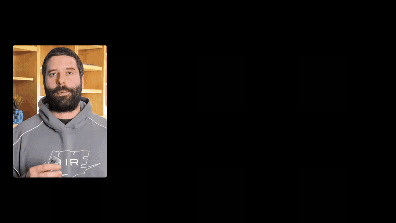 The image shows a man with a beard and mustache, wearing a gray hoodie with the Nike logo, standing in front of a wooden bookshelf. He is holding a small, dark-colored object in his right hand. The man's gaze is directed towards the camera, creating a sense of engagement with the viewer. The background is a simple blac